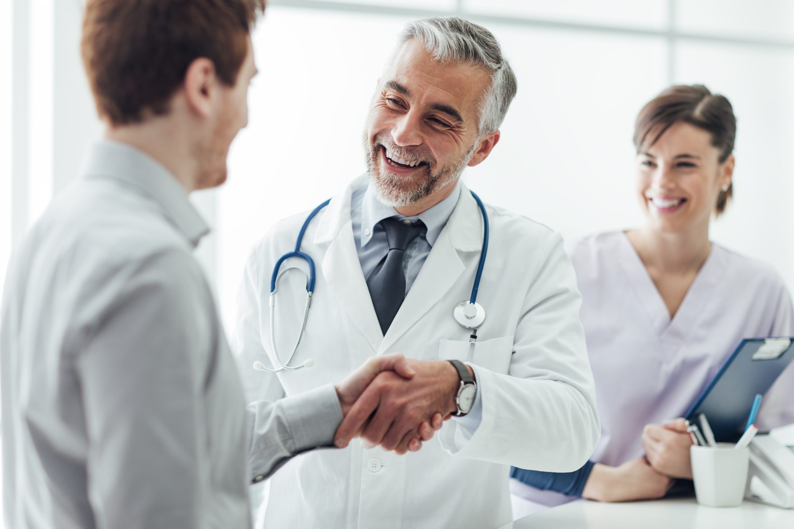 Ein Arzt und ein Patient schütteln sich die Hände, während eine Pflegekraft im Hintergrund lächelt.