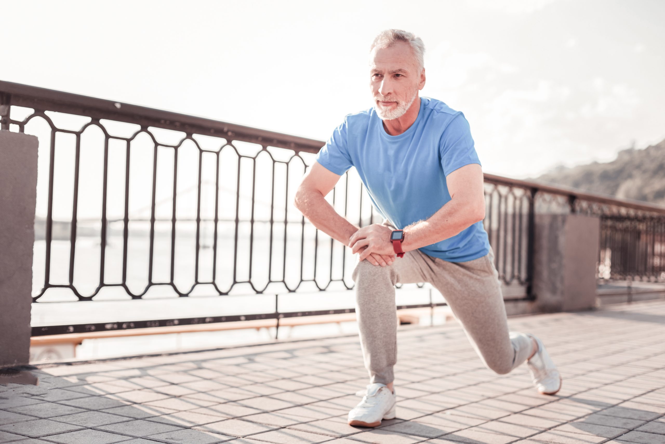 Älterer Mann dehnt sich auf einer Promenade, trägt blaues T-Shirt und graue Jogginghose.