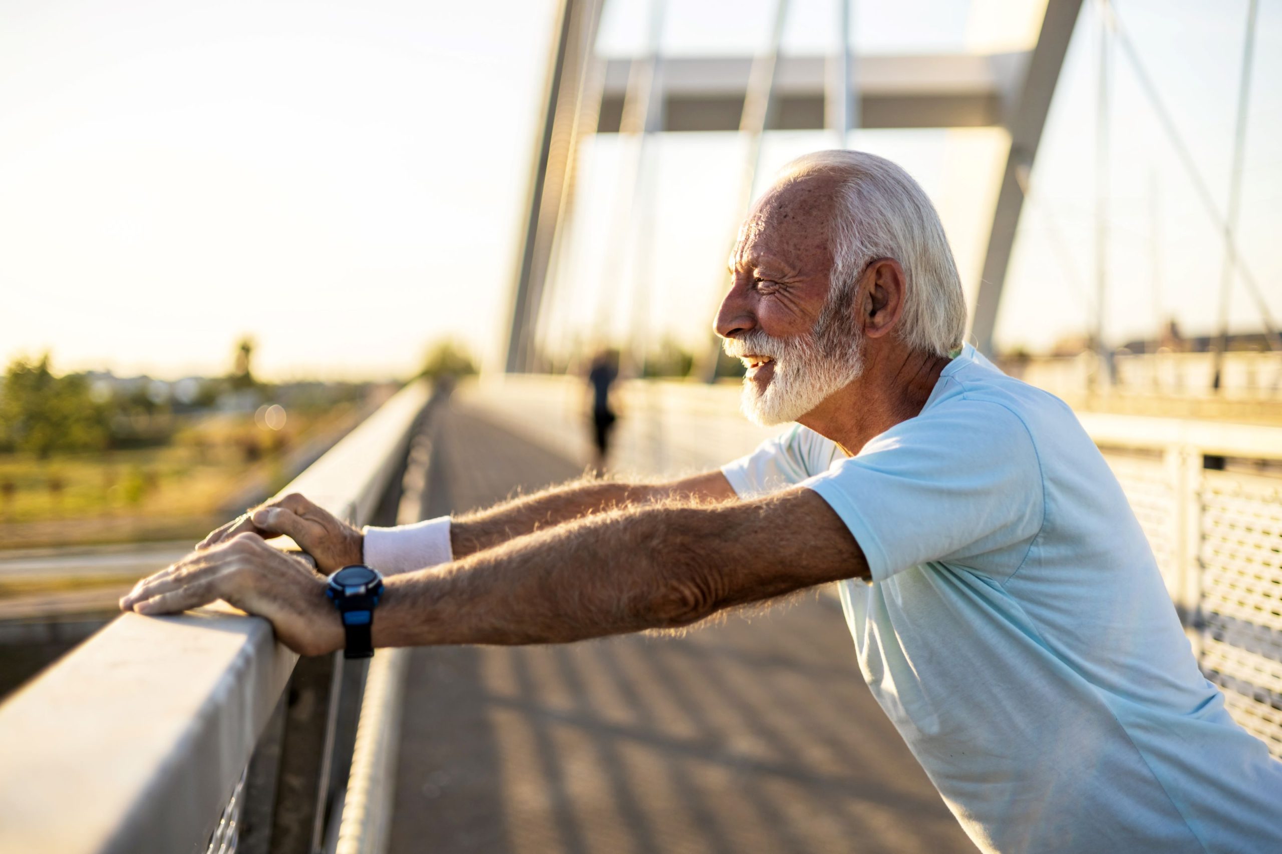 Älterer Mann streckt sich lächelnd auf einer Brücke im Sonnenlicht.