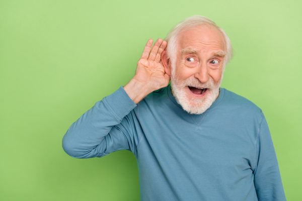 Ein älterer Mann in blauem Pullover hält seine Hand an sein Ohr, vor grünem Hintergrund.
