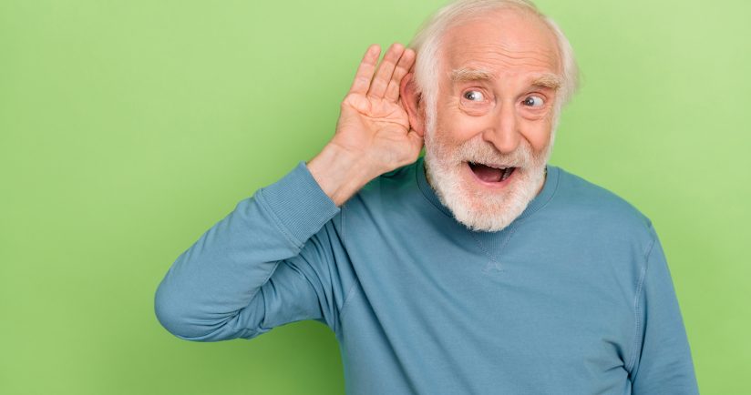 Ein älterer Mann in blauem Pullover hält seine Hand an sein Ohr, vor grünem Hintergrund.