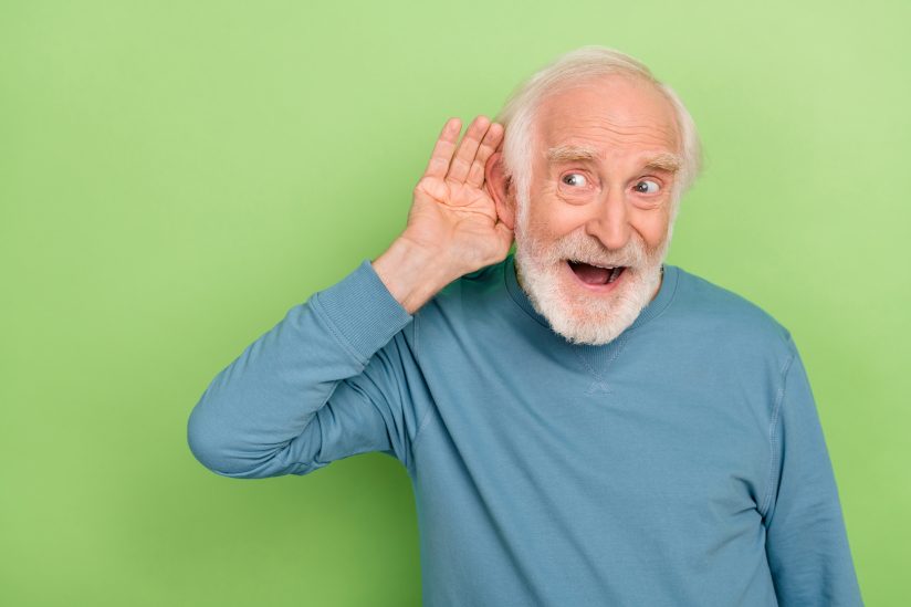 Ein älterer Mann in blauem Pullover hält seine Hand an sein Ohr, vor grünem Hintergrund.