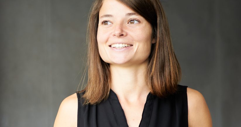 Eine lächelnde Person mit schulterlangem Haar in schwarzer Kleidung vor grauem Hintergrund.