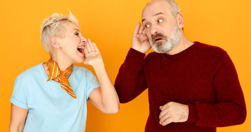 Eine Frau flüstert einem Mann zu. Beide stehen vor einem orangefarbenen Hintergrund.