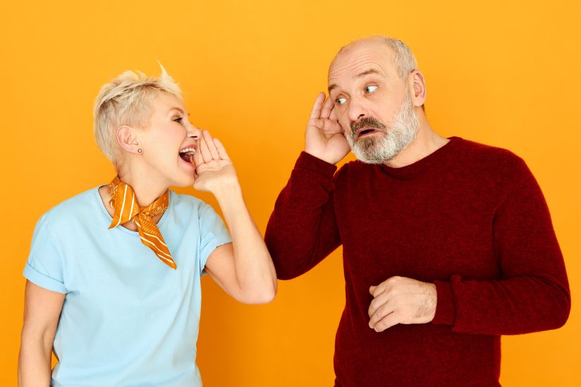 Eine Frau flüstert einem Mann zu. Beide stehen vor einem orangefarbenen Hintergrund.