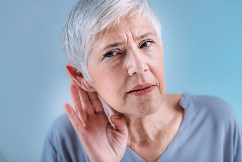 Eine ältere Person mit weißen Haaren hält eine Hand ans Ohr. Sie schaut konzentriert. Hintergrund in Blau.